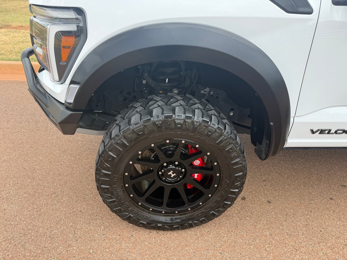 Close-up of a white pickup truck's all-terrain tire and custom black wheel with red brake calipers on gravel